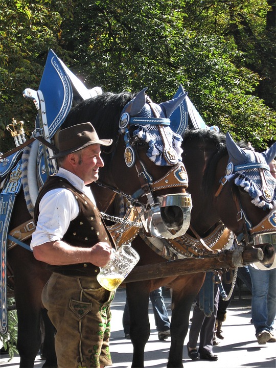 Photo of a man in traditional Bavarian dress holding a beer next to horses in armour